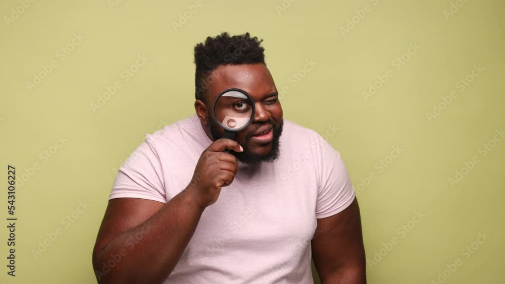 Funny African-american man wearing pink t-shirt holding magnifying glass and looking around with big zoom eye, positive face. Indoor studio shot isolated on light green background. Funny African-american man wearing pink t-shirt holding magnifying glass and looking around with big zoom eye, positive face. Indoor studio shot isolated on light green background.
