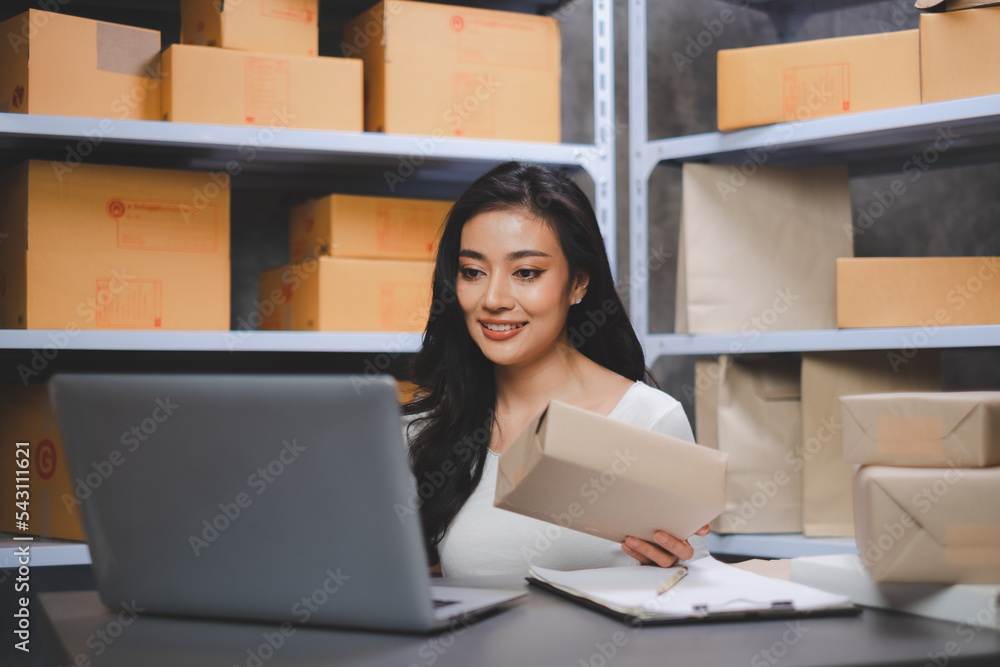 Asian Start-up businesswoman is smiling and cheking parcel boxes with checklist in laptop for delivery the product. Entrepreneur SME working with box at home. Concept of online marketing at home. Asian Start-up businesswoman is smiling and cheking parcel boxes with checklist in laptop for delivery the product. Entrepreneur SME working with box at home. Concept of online marketing at home.