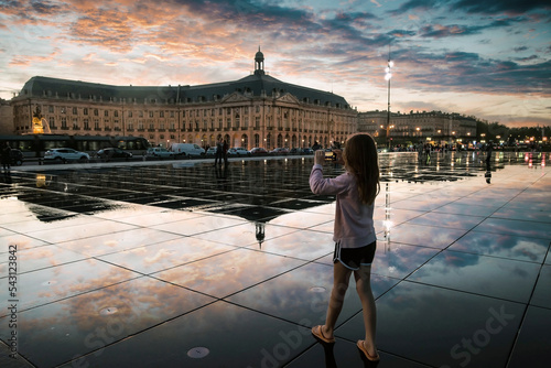 Young girl taking a photo of the famous Bordeaux water mirror with Place de la Bourse in the background at sunset photo