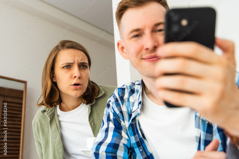 Jealous woman reads her husband's message on the phone, standing behind him, while he texts, sitting in the kitchen. Infidelity and deceit, the problem of jealousy in a relationship. selective focus Jealous woman reads her husband's message on the phone, standing behind him, while he texts, sitting in the kitchen. Infidelity and deceit, the problem of jealousy in a relationship. selective focus