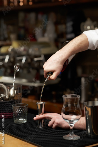 Cropped close-up shot of a bartender is pouring drink into a stainless steel jigger. Men s hands are pouring drink into a measuring cup on the bar counter. Front view.