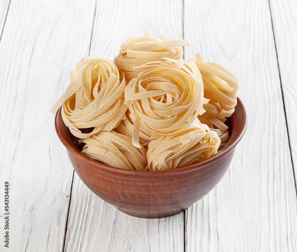 Uncooked tagliatelle pasta in ceramic bowl on white wooden background
