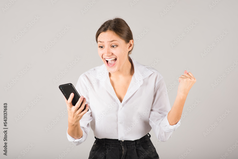 The businesswoman shows a victory gesture, shouts and uses a mobile phone on a gray background. The businesswoman shows a victory gesture, shouts and uses a mobile phone on a gray background.