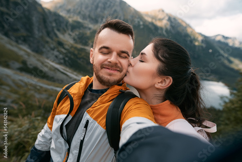 Happy family of man and woman spending vacation time in the mountain. Young couple kissing on a cheek with a lake and mountain background.
