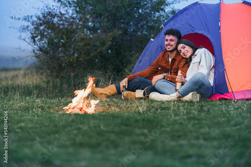 A happy couple is sitting and talking in front of a tent by the fire while enjoying the sunset. They enjoy camping in nature.