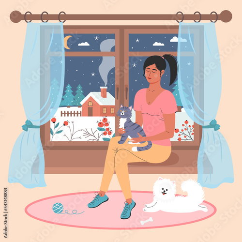 Girl with a cat sitting on the window. White spitz lying on the rug nearby. There is a winter night landscape outside the window. Winter mood, love pets, relaxing, cozy home concept.