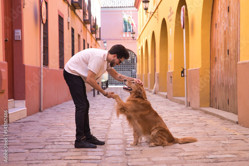 Young Hispanic man with beard, sunglasses and white shirt, holding his paw and petting his dog on a lonely street. Concept animals, dogs, love, pets, golden.