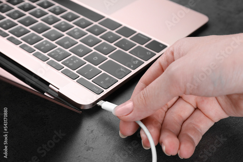 Woman plugging USB cable with lightning connector into laptop port on dark table, closeup photo