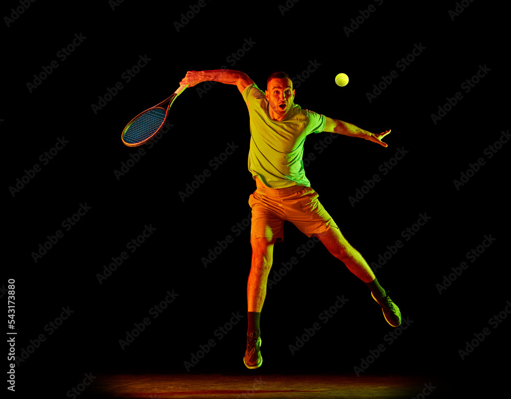 Dynamic portrait of energetic male tennis player in sportwear playing tennis isolated over dark background in neon light. Concept of motion, speed, professional sport.