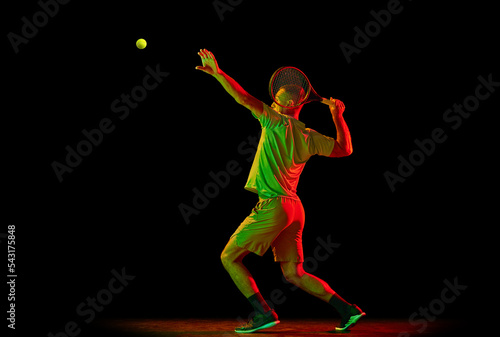 Dynamic portrait of energetic male tennis player in sportwear playing tennis isolated over dark background in neon light. Concept of motion, speed, professional sport.