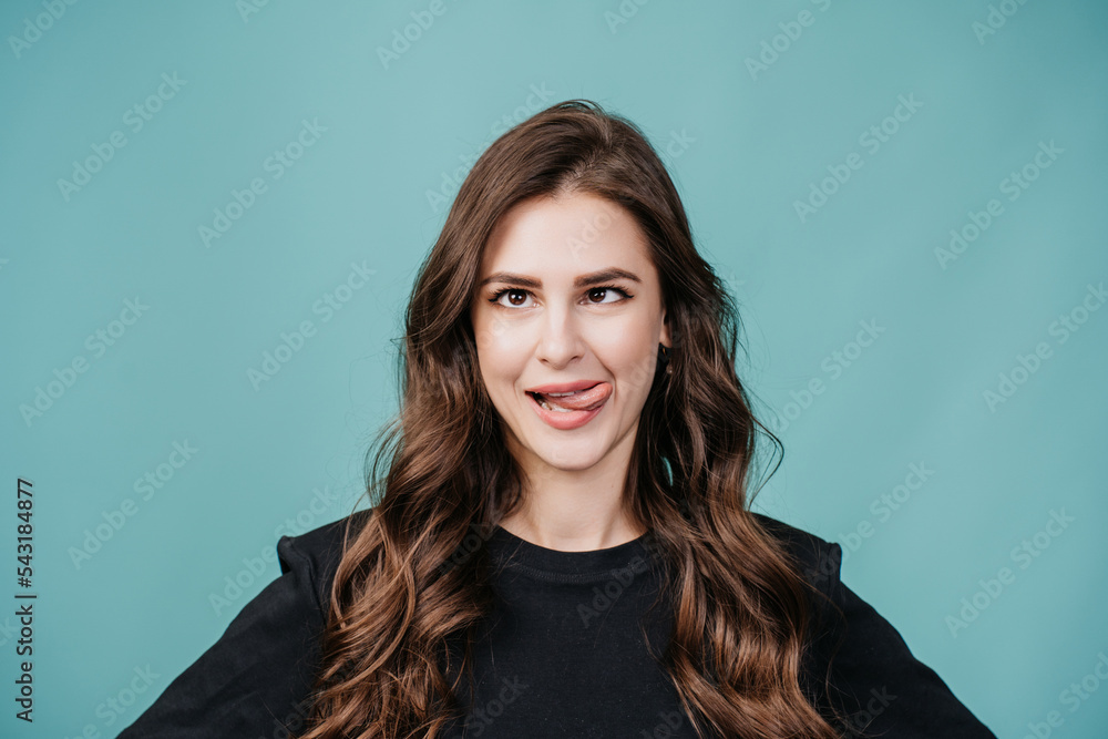 Fototapeta premium Playful brunette young woman in black sweater shows tongue, squinted eyes over turquoise background at studio. Close up portrait of hispanic girl with mad face expression. Having fun, joking. Drunk