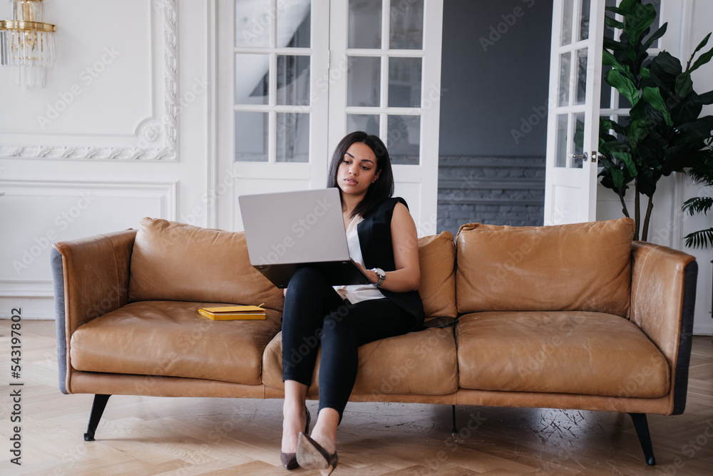 Overloaded Brazilian businesswoman working home sitting on cozy leather sofa using laptop. Tired African American entrepreneur making video call. Beautiful hispanic woman remote studying. Education.