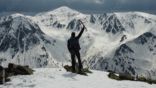 Alpinist on the top of the snowy mountain with his back, raised arm holding an ice tool