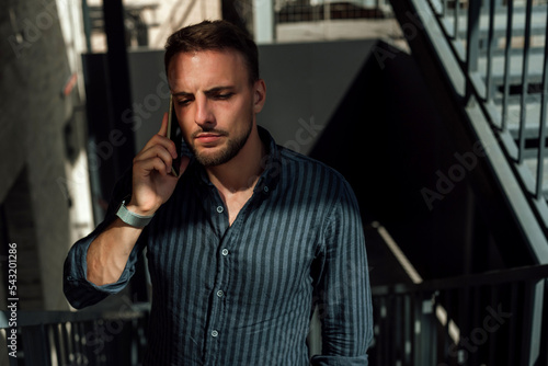 Wearing a dark blue striped shirt; street male portrait; a guy with a watch on his hand is talking on the phone and standing on the stairs