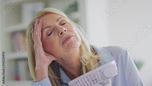 Menopausal mature woman at home having hot flush and cooling herself with electric fan shot in slow motion photo