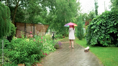 a girl walks in the rain barefoot along the path of the summer garden with a funny multi-colored umbrella. High quality 4k footage
