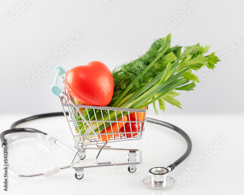 Health care still life with organic vegetables in a shopping cart with decorative heart and stethoscope on a white table. Concept of healthy food and control of good health with a diet