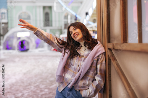 Funny young caucasian woman streches hand away smiling keeps hand in pocket. Brunette wears jeans, shirt and sweater. Concept of positive moments. photo