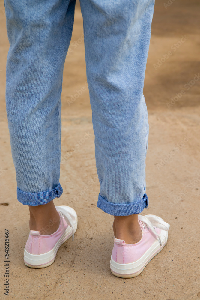 Side view of the legs of a teenage girl in pink sneakers and blue jeans against a background of yellow dry grass. The concept of clothing for modern teenagers in the cool season. Vertical photo