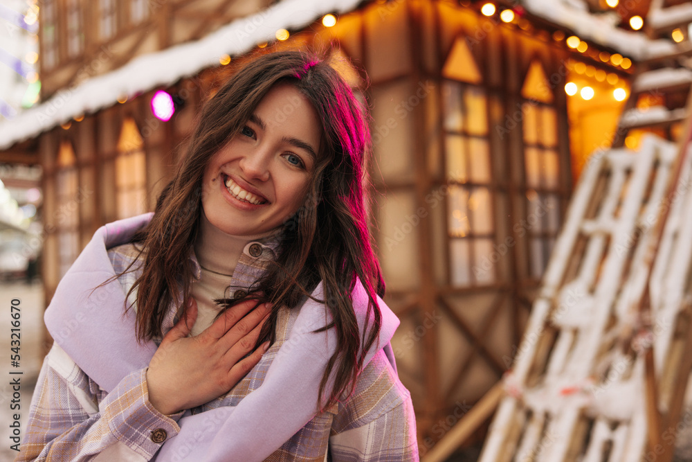 Cheerful young caucasian girl looking at camera with smile walks in park during winter holidays. Brunette wears shirt and sweater. Leisure lifestyle and beauty concept. Cheerful young caucasian girl looking at camera with smile walks in park during winter holidays. Brunette wears shirt and sweater. Leisure lifestyle and beauty concept.