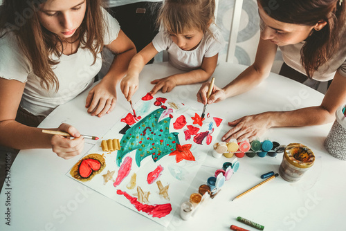 family, children, girls, sisters draw a Christmas tree at home on the table with their mother, daughters stained their mother with paints, she smiles