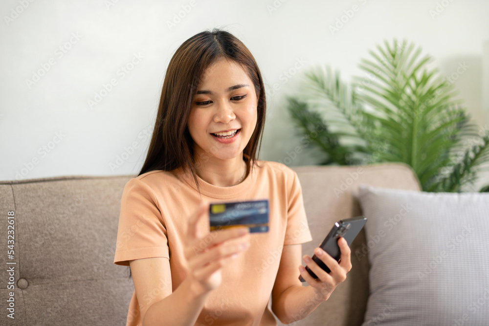 Young asian woman is holding new credit card and smartphone while sitting on comfortable sofa and entering Young asian woman is holding new credit card and smartphone while sitting on comfortable sofa and entering