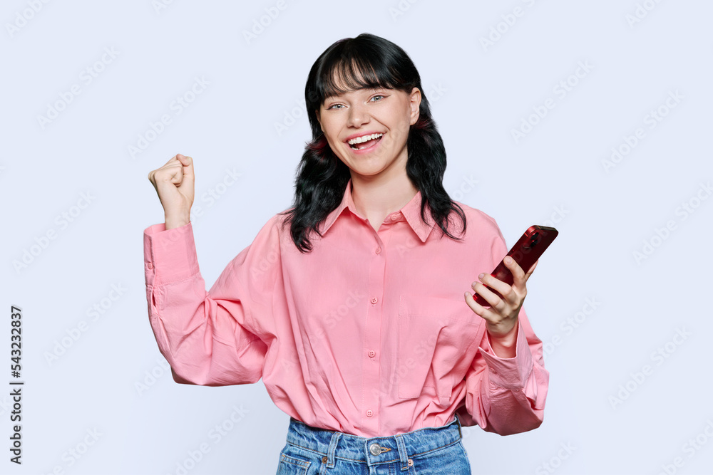 Teenage female student with smartphone making yes gesture on light background Teenage female student with smartphone making yes gesture on light background