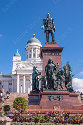 Helsinki, Finland - July 19, 2022: Statue of Tsar Alexander II of Russia on Senate Square with white cathedral behind under blue sky. Greenish bronze statue combinations and flowers photo