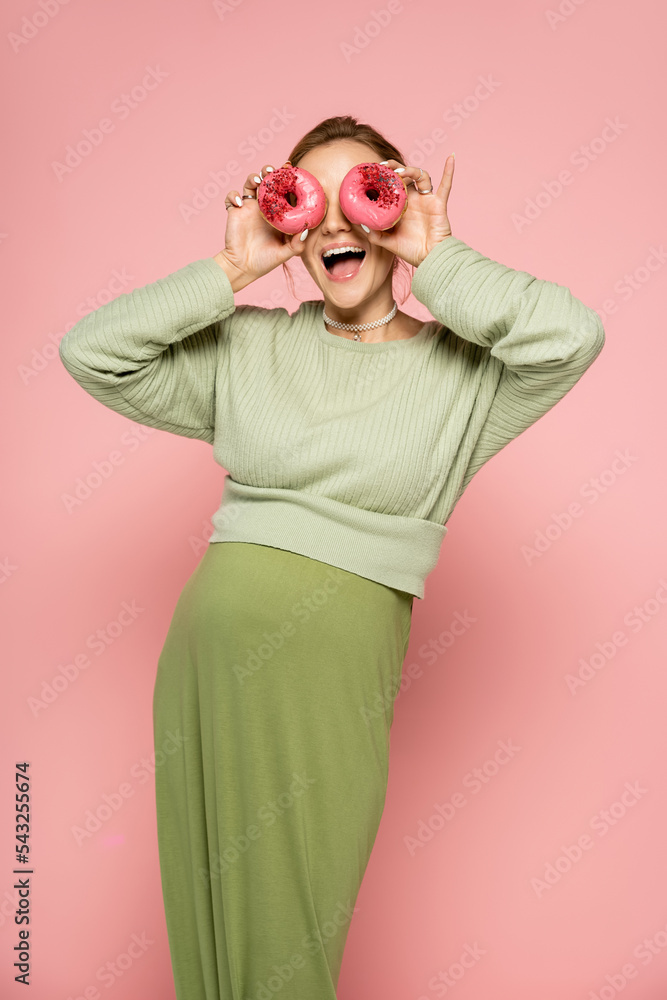Excited pregnant woman covering face with donuts on pink background. Excited pregnant woman covering face with donuts on pink background.