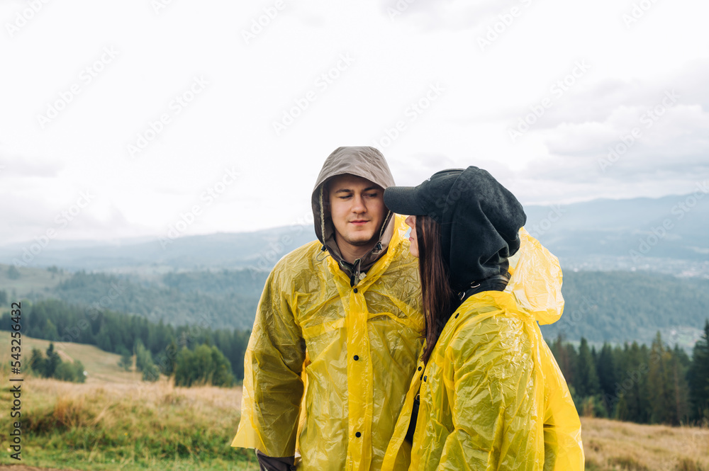 A beautiful young couple on a hike stands in the mountains against the background of beautiful views, dressed in raincoats and looking at each other with a serious face.