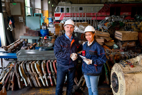 Caucasian factory worker or technician stand and shake hands with Asian factory woman in front of the train and spare part of machine in workplace area also look at camera.