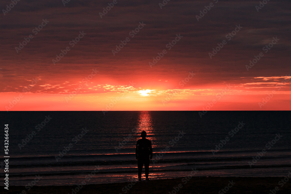 The shore of the Caspian Sea at sunset, pink-orange clouds, water, beach, summer, a person stands on the shore. Silhouette of a man. The shore of the Caspian Sea at sunset, pink-orange clouds, water, beach, summer, a person stands on the shore. Silhouette of a man.