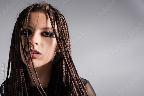 portrait of woman with braids and stylish makeup with piercing looking at camera isolated on grey.