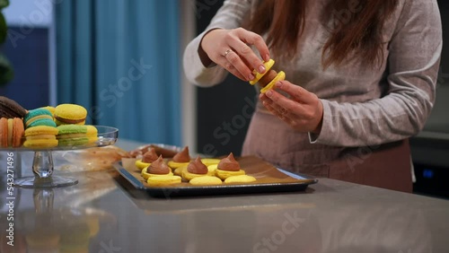 Unrecognizable Caucasian plus-size lady making french macaroni with chocolate icing in kitchen in slow motion. Young woman cooking yellow cookies with caramel buttercream indoors photo