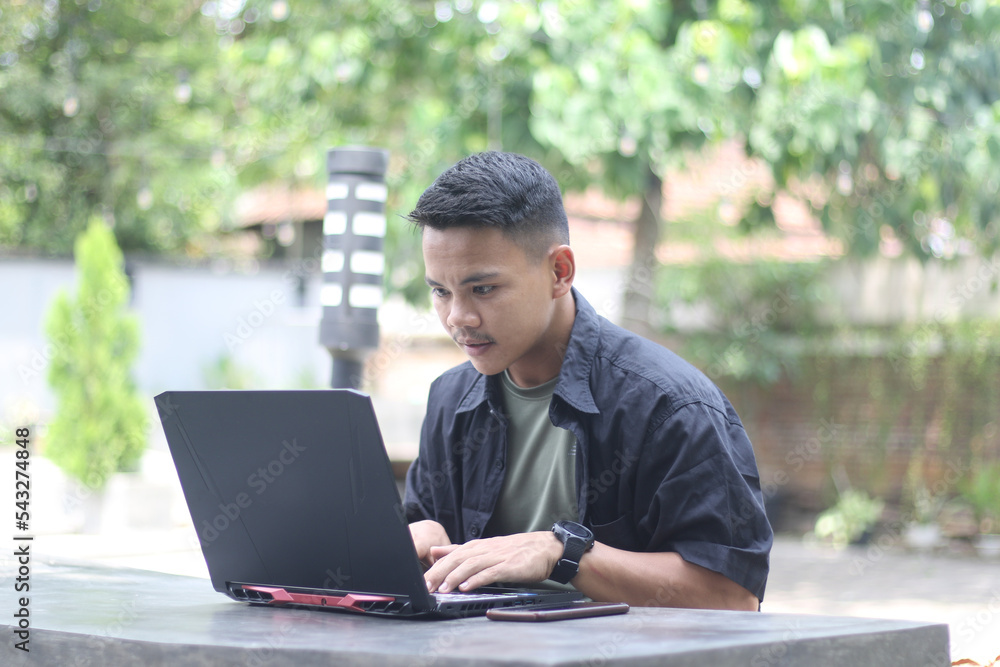 Attractive young Asian man using laptop in co-working space with happy face Attractive young Asian man using laptop in co-working space with happy face