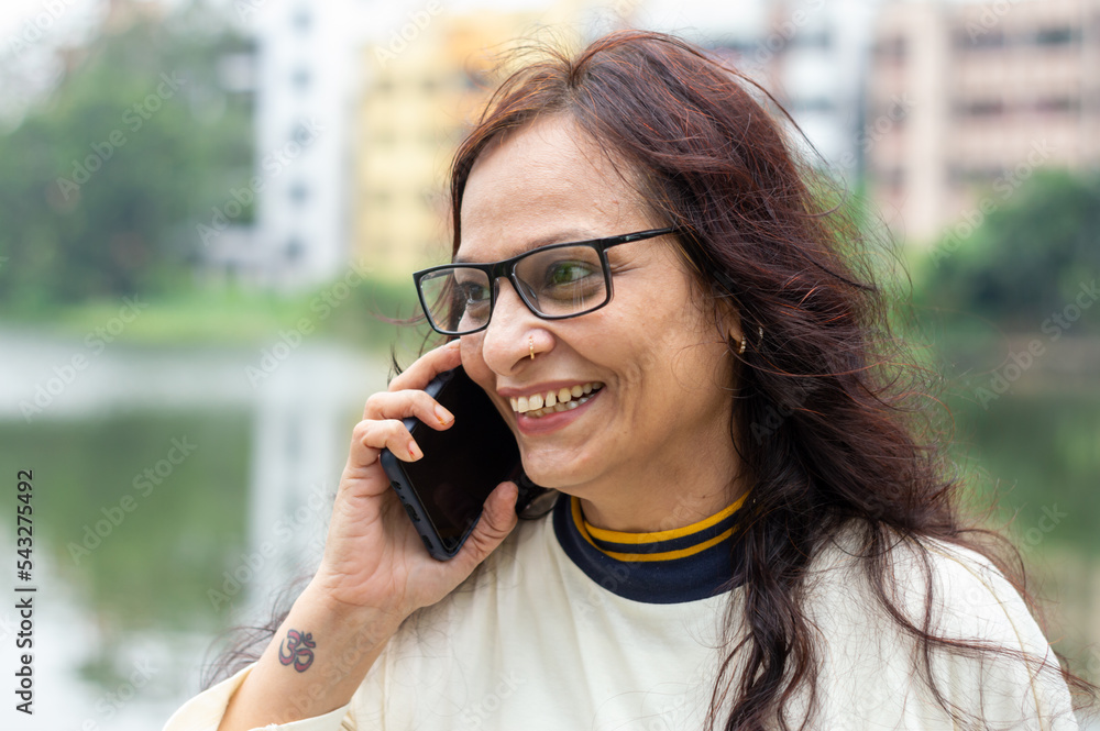Mid Adult Smiling Woman with long brown hair Speaking on mobile phone outdoors. Closeup. She is wearing White T-shirt and specs. Front view. Head and Shoulder Shot. Garden lake side in the background. Mid Adult Smiling Woman with long brown hair Speaking on mobile phone outdoors. Closeup. She is wearing White T-shirt and specs. Front view. Head and Shoulder Shot. Garden lake side in the background.