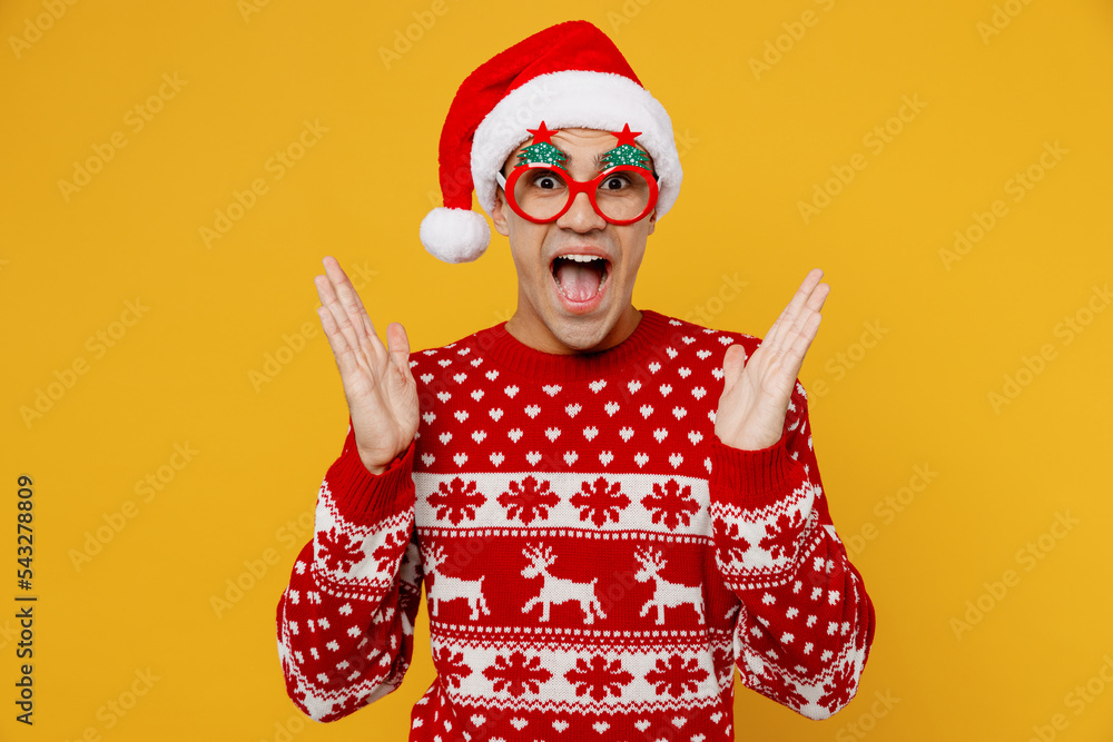 Merry shocked fun young man wear red knitted sweater Santa hat Christmas glasses look camera spread hands posing isolated on plain yellow background. Happy New Year 2023 celebration holiday concept.