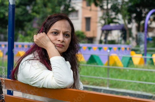 Mid Adult worried Woman sitting on hand on chin pose in a park bench and looking at a distance. Front view. Indian Ethnicity Age 50 to 54 Years. photo