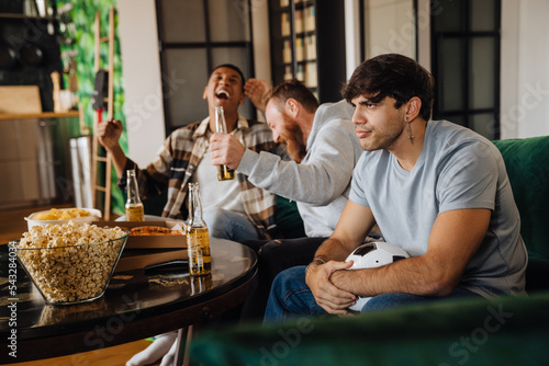 Displeased man watching sports match while his friends celebrating victory in living room