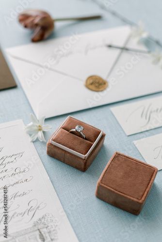 Engagement ring with a diamond in a box on a light blue background. Invitations on handmade paper with calligraphy and wax seal.