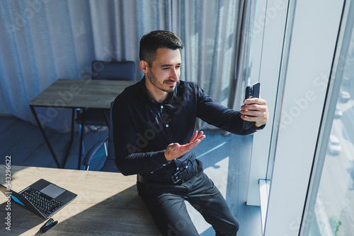 Young successful male worker in glasses and black shirt sitting on desk and working on project via laptop in modern office looking at screen