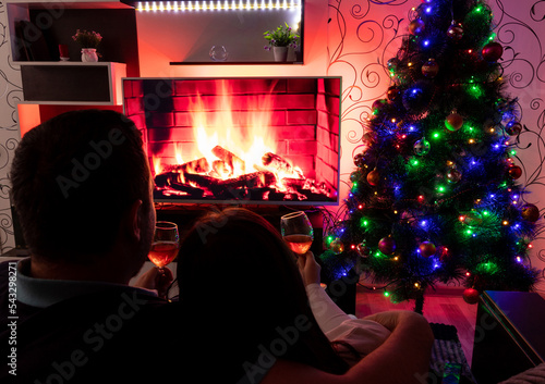 Couple hugs and sits on sofa, holding glass of wine in front of TV, which depicts fireplace. Selective focus. Images for articles about leisure, winter, family