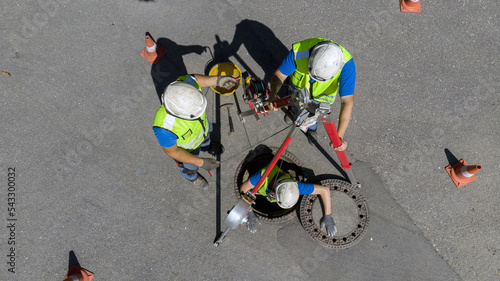 Maintenance and washing of a cesspool through the access of a road manhole. Skilled workers at work lower themselves underground using a pulley