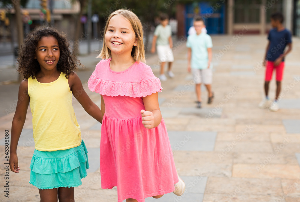 Happy blonde preteen girl walking with her african american girl playmate along city street on summer day ..