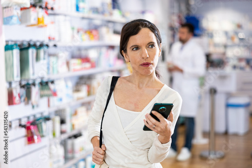 Woman chooses medical products in pharmacy, checking the list on a smartphone