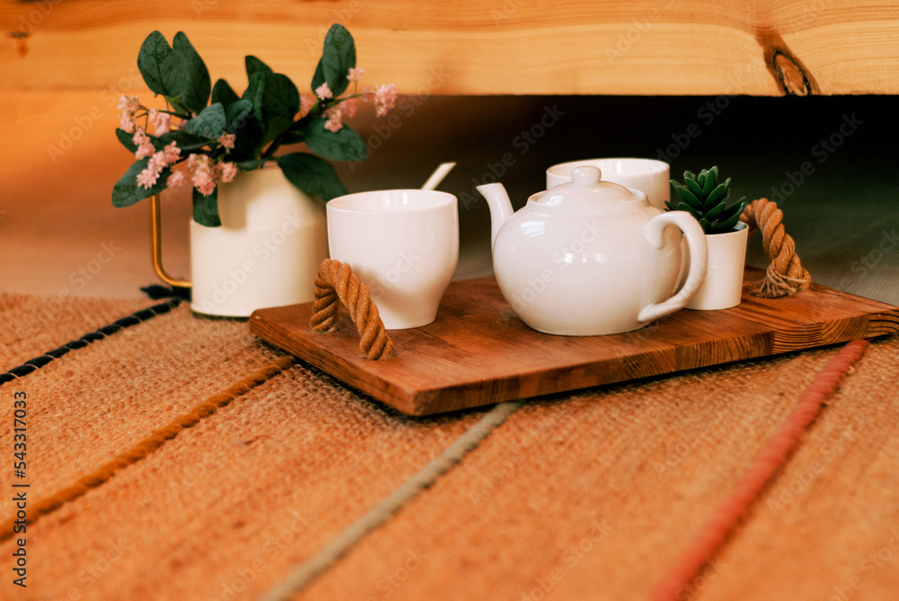 Tea ceremony. White porcelain cups and a sugar bowl on a wooden tray. Porcelain vase with flowers. Scandinavian interior with elements of textiles and wooden decor. Home comfort. Wooden flooring