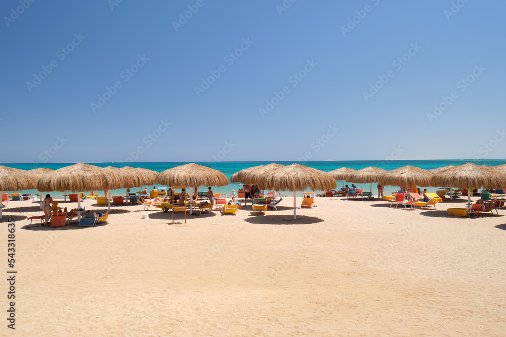 Straw shade umbrellas on sea tropical beach with resting sunbeds against blue vibrant sky in summer