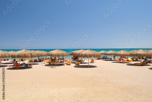 Straw shade umbrellas on sea tropical beach with resting sunbeds against blue vibrant sky in summer