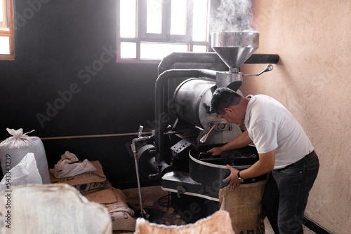 An Hispanic man is picking up the coffee with a sack from the roaster machine