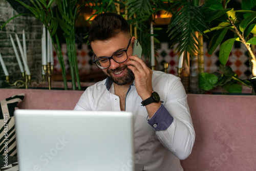 Portrait of smiling man with beard sitting with laptop, talking on phone photo